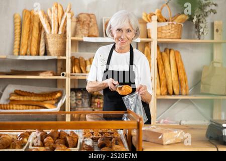 Die Senior-Verkäuferin bringt Croissant in eine Plastiktüte für den Käufer, verpackt Waren. Stockfoto