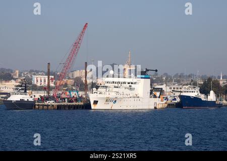 San Diego, Kalifornien, USA. Dezember 2024. Das markante weiße Schiff im Zentrum ist ein NOAA-Forschungsschiff, das Teil der Flotte der National Oceanic and Atmospheric Administration ist und für ozeanographische und atmosphärische Forschungsmissionen entwickelt wurde. Das Schiff ist mit modernster Technologie ausgestattet und unterstützt die Datenerfassung, die für das Verständnis von Meeres- und Klimasystemen von entscheidender Bedeutung ist. Daneben unterstreichen andere angedockte Schiffe und Industriekräne den dynamischen Betrieb im Hafen von San Diego und zeigen die maritime und wissenschaftliche Bedeutung des Gebiets. (Foto: © Ian L. Sitren/ZUMA Press Wire) EDITORI Stockfoto