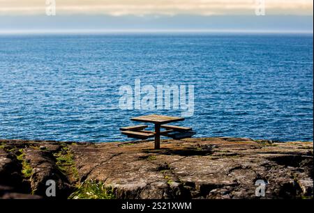 Ein verwitterter hölzerner Picknicktisch auf einer felsigen Klippe mit Blick auf das weite Meer, unter einem blauen Himmel mit hellen Wolken und fernen Berglandschaften Stockfoto