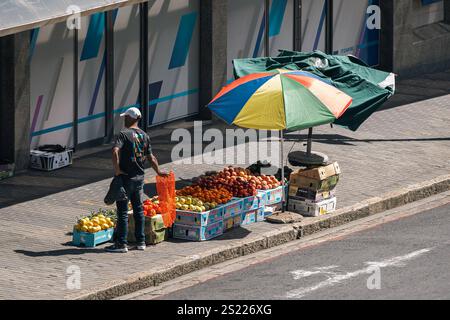 Der Mann verkauft Obst aus Kisten auf der Straße auf einem Fußgängerweg mit Kopfsteinpflaster neben der Straße. Stadtbild. Kapstadt, Südafrika - 31. Oktober 2023 Stockfoto