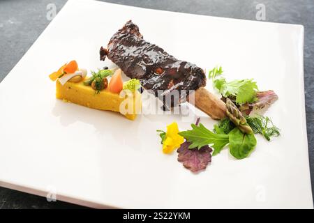 Elegantes Gourmet-Gericht aus glasierten kurzen Rippchen mit Polenta, Gemüse und Wildkräutersalat auf einem weißen Teller, Hauptgericht in einem Gourmet-Menü, Kopierraum, s Stockfoto