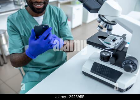 Männlicher Chemiker mit Smartphone für Online-Forschung bei der Analyse von Proben unter dem Mikroskop Stockfoto