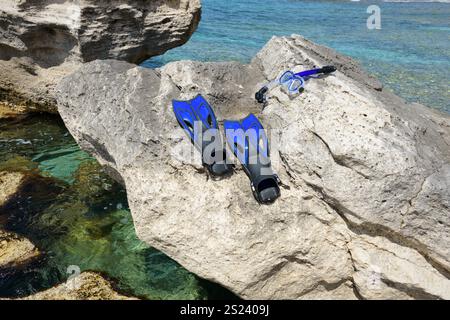 Ein Paar blaue Schnorchelflossen und eine Maske werden lässig auf einem sonnendurchfluteten Felsen an der Küste platziert, wobei klares türkisfarbenes Wasser sanft auf die Felsen schlägt. Stockfoto