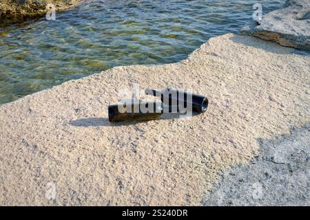 Zwei schwarze Flaschen stehen auf sonnendurchfluteten Felsen neben klarem Wasser, was eine ruhige Atmosphäre am Ufer an einem warmen Nachmittag schafft. Stockfoto