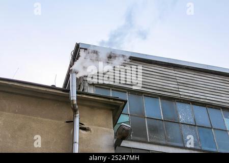 Weißer Rauch steigt aus einem Auspuffrohr eines Industriegebäudes bei klarem Himmel auf, was auf eine mögliche Herstellungstätigkeit oder Heizung hinweist. Stockfoto