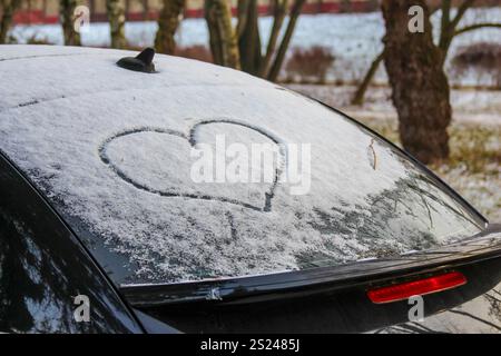 Eine Herzform ist in den Schnee eingeätzt, der ein geparktes Auto bedeckt und vor einem Hintergrund von Bäumen in einer winterlichen Umgebung liegt. Stockfoto