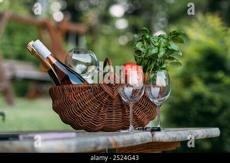 Rustikaler Korb mit Wein, Gläsern, Basilikum und Tomaten auf einem Holztisch in ruhiger Atmosphäre im Freien. Farm-to-Table-Wohnkonzept. Stockfoto