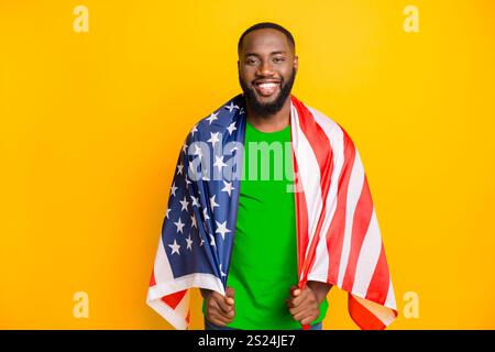Foto eines schwarzen amerikanischen Patrioten, der die amerikanische Flagge aufgesetzt hat, während er isoliert steht, mit gelbem Hintergrund Stockfoto