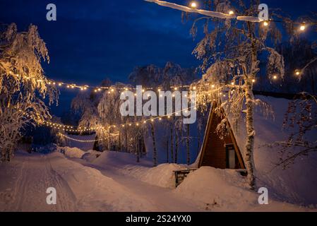 Schneebedeckte Holzhütten mit A-Rahmen schaffen ein magisches Winterwunderland, das mit warmen Lichtern unter ruhigen Bäumen in einer friedlichen Schneelandschaft leuchtet Stockfoto