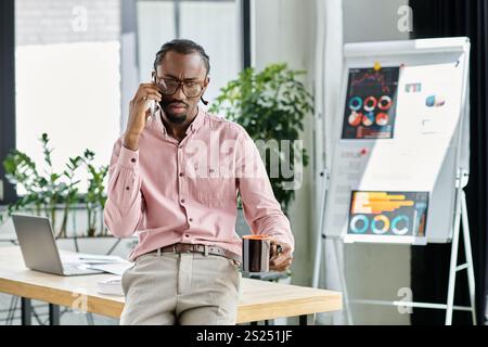 Ein fokussierter junger afroamerikanischer Mann, der während eines Anrufs mehrere Aufgaben erledigt, während er an einem schicken Arbeitsplatz Kaffee schlürft. Stockfoto