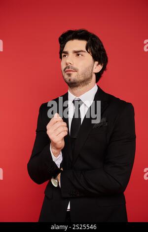 Ein brünette Mann steht selbstbewusst in einem schwarzen Anzug und denkt über das Leben vor einer kühnen roten Wand nach. Stockfoto