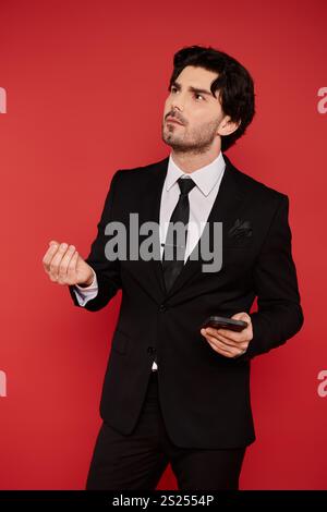 Brünette Mann in einem stilvollen Anzug schaut nachdenklich nach oben und hält ein Telefon in einem hellen Raum. Stockfoto