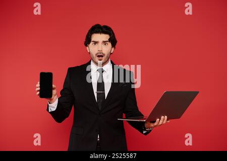 Ein junger Mann zeigt einen Ausdruck der Überraschung, während er ein Notebook und ein Smartphone zusammenhält. Stockfoto
