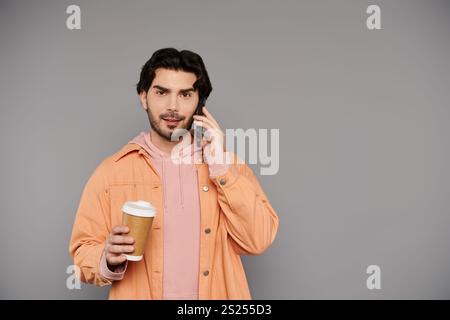 Ein junger Mann mit braunen Haaren führt ein Telefongespräch, während er eine warme Kaffeetasse hält. Stockfoto