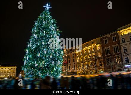 Ein großer, hell geschmückter Weihnachtsbaum steht nachts auf einem geschäftigen Stadtplatz, der wunderschön inmitten einer festlichen Menschenmenge und umgeben von seinem leuchtet Stockfoto