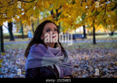 Eine lächelnde Frau in einen Lavendelschal gewickelt, die goldene Blätter in einem ruhigen Herbstpark ansieht. Warme Saisontöne und traumhafte Atmosphäre. Stockfoto