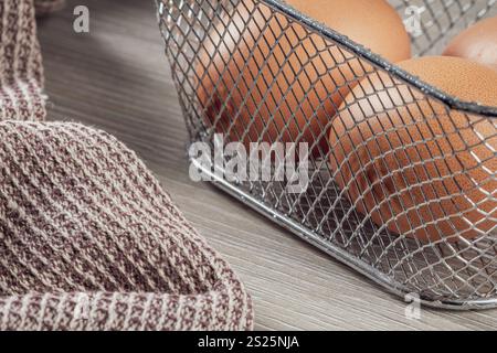 Hühnereier in Metallkorb und Leinenserviette auf Küchentisch, Vorderansicht aus nächster Nähe. Stockfoto