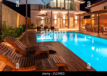 Luxusvilla mit beleuchtetem Swimmingpool und Holzliegen bei Nacht. Konzept der modernen Hinterhofentspannung Stockfoto