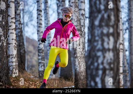 Sportliche Läuferin in rosafarbener Jacke und gelber Jogginghose, die auf einem Wanderweg im Wald läuft Stockfoto