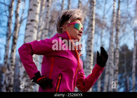 Sportliche Läuferin in rosafarbener Jacke und gelber Jogginghose, die auf einem Wanderweg im Wald läuft Stockfoto