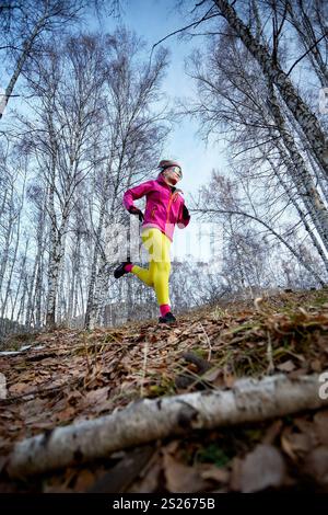 Sportliche Läuferin in rosafarbener Jacke und gelber Jogginghose, die auf einem Wanderweg im Wald läuft Stockfoto