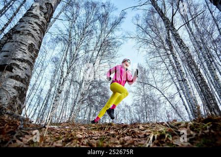 Sportliche Läuferin in rosafarbener Jacke und gelber Jogginghose, die auf einem Wanderweg im Wald läuft Stockfoto