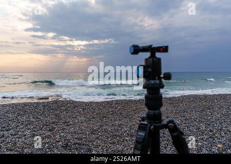 Eine ruhige Strandszene bei Sonnenaufgang mit einem Kamerastativ an einem Kieselstrand, das die friedlichen Wellen und den weiten Himmel einfängt. Ideal für Fotokonzepte Stockfoto