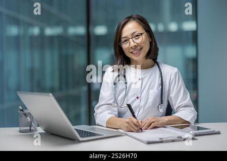Porträt einer jungen asiatischen Ärztin, einer lächelnden Ärztin, die in die Kamera blickt, mit Dokumenten und Papieren arbeitet, die im Büro am Arbeitsplatz arbeiten. Stockfoto