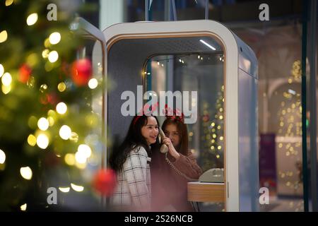 Zwei Freunde mit Kopfbügeln im Urlaubsstil genießen einen fröhlichen Moment in einer modernen Telefonzelle. Die Einstellung wird durch warme Lichter, cr, aktiviert Stockfoto