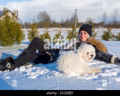 Fröhliche Frau in schwarzer Winterkleidung, die auf klarem Schnee liegt und während der Sonne spielerisch mit flauschigem weißem Bichon-Frise im umzäunten Hinterhof interagiert Stockfoto