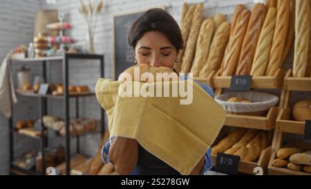 Frau genießt das Aroma von frisch gebackenem Brot in einer Bäckerei, die von Regalen mit verschiedenen Broten und Gebäck umgeben ist. Stockfoto