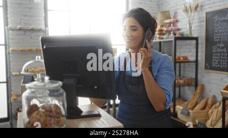 Frau, die in der Bäckerei telefoniert, trägt eine Schürze hinter der Theke mit Gebäck und Brot im Hintergrund Stockfoto