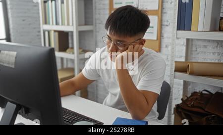 Junger Mann, der am Schreibtisch im Heimbüro arbeitet, sieht müde und gelangweilt aus, mit Computer, Büchern und Regalen im Hintergrund Stockfoto