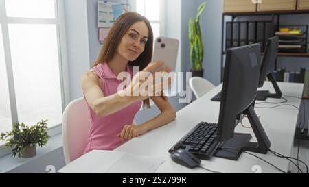 Junge Frau, die Selfie in moderner Büroumgebung mit Desktop-Computer und Innenanlagen macht, mit einer professionellen Arbeitsumgebung Stockfoto