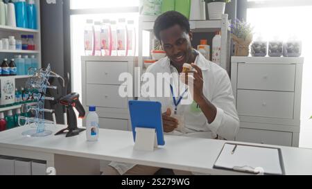 Junger Mann in einer Apotheke zeigt eine Flasche Pillen während eines Videogesprächs, umgeben von verschiedenen Store-Produkten und medizinischen Geräten auf einem gut organisierten Stockfoto