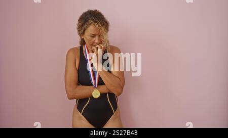 Frau, die auf rosafarbenem Hintergrund im Badeanzug mit Goldmedaille und nachdenklichem Ausdruck steht Stockfoto
