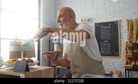 Senior Mann mit grauen Haaren und Bart, der Gebäck in einer Bäckerei mit Brotregalen im Hintergrund serviert. Stockfoto