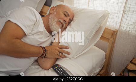 Mann mit grauen Haaren und Bart, der zu Hause friedlich im Bett schläft, bietet ein ruhiges und komfortables Innenschlafzimmer mit natürlichem Lichtfilter Stockfoto