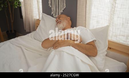 Ein älterer Mann mit grauen Haaren und Bart schläft friedlich in seinem Bett in einem gemütlichen Schlafzimmer, mit Innenbeleuchtung und Inneneinrichtung. Stockfoto