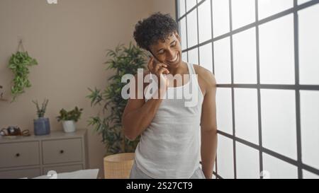 Junger Mann, der am Schlafzimmerfenster steht und telefoniert, gekleidet in einem weißen Tank, mit Topfpflanzen im Innenbereich, hellem Tageslicht und einem gemütlichen M Stockfoto