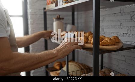 Mann, der frisch gebackene Croissants auf einer Holzplatte in einer Bäckerei mit weißen Ziegelwänden und Regalen mit verschiedenen Brotauflagen platziert, indo Stockfoto