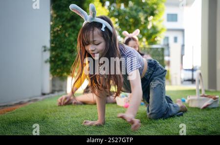 Zwei Kinder nehmen an einer Ostereierjagd in einem lebhaften Hinterhof Teil. Ein Kind krabbelt mit Hasenohren auf dem Gras, während das andere nahe erforscht Stockfoto