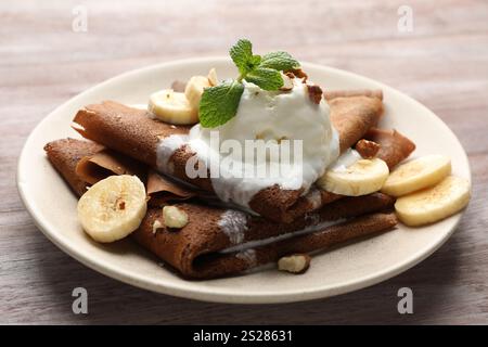 Köstliche Schokoladencrepes mit Banane, Walnüssen, Minze und Eislöffel auf Holztisch, Nahaufnahme Stockfoto