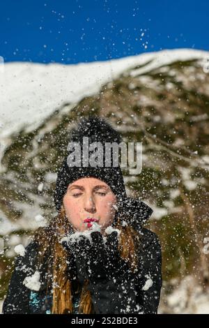 Eine junge Frau, die Schnee aus ihren Händen bläst und eine Menge Schneeflocken in einem Winterwunderland erzeugt. Stockfoto