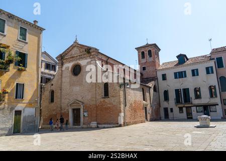 Eine historische Backsteinkirche mit einem Glockenturm auf einem ruhigen venezianischen Platz, umgeben von bunten Gebäuden mit verschlossenen Fenstern unter klarem Himmel. Stockfoto