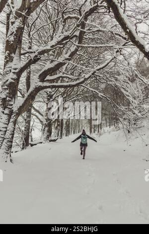 Eine Wanderer mit blauem Rucksack, die an einem atemberaubenden Wintertag in der Nähe von Skipton in North Yorkshire durch schweren Schnee unter einer Allee mit Bäumen spaziert. Stockfoto