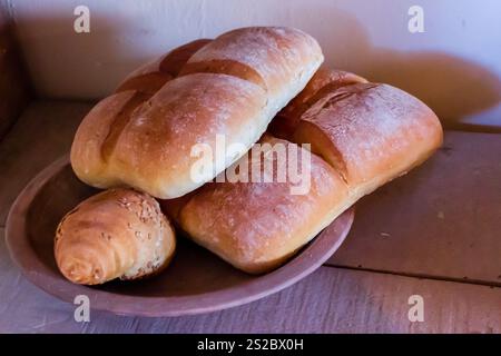 Eine Sammlung von weichen, frisch gebackenen Brötchen, einschließlich einer Sesambrötchen, serviert auf einer Keramikplatte mit warmer Beleuchtung vor rustikaler Kulisse, Stockfoto