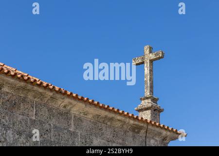 Religiöses christentum-Kreuz aus Granit auf dem Dach einer Kirche mit blauem Himmel in Quintela Portugal. Stockfoto