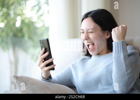 Aufgeregte asiatische Frau, die Erfolg feiert und das Telefon überprüft, das zu Hause auf einer Couch sitzt Stockfoto