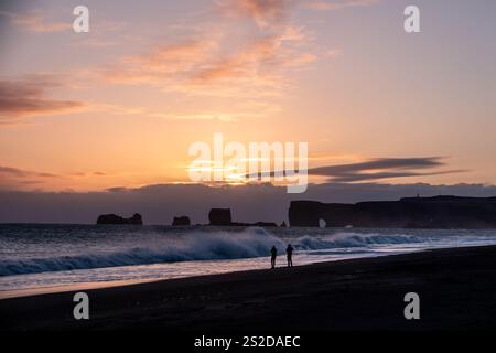 Entfernte Silhouette von zwei Personen, die am schwarzen Sandstrand von Reynisfjara bei Sonnenuntergang entlang laufen, Süd-Zentral-Island, Island Stockfoto